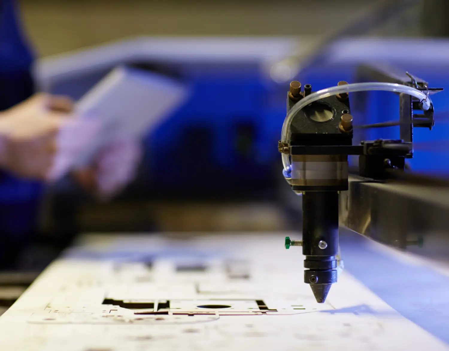 A close-up of a precision cutting tool, poised above a detailed design on a sheet, with a person holding a tablet in the background A close-up of a precision cutting tool, poised above a detailed design on a sheet, with a person holding a tablet in the background
